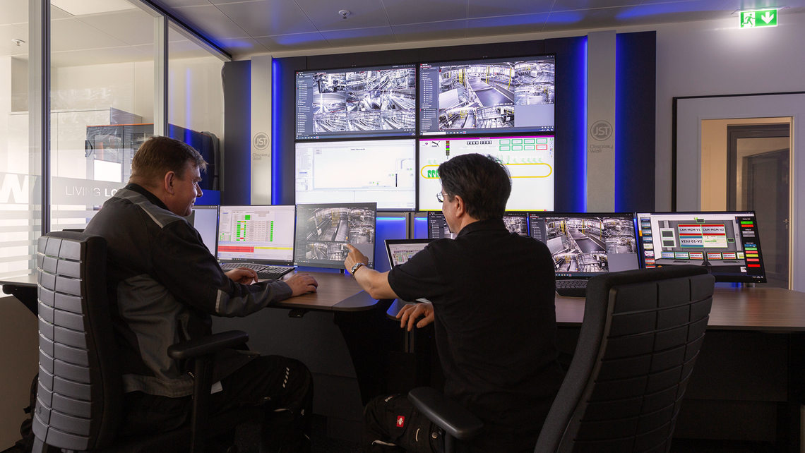 Two men at a shared desk looking through computer screens and large wall monitors, keeping track of operational excellence through TGW Logistics' warehouse management software