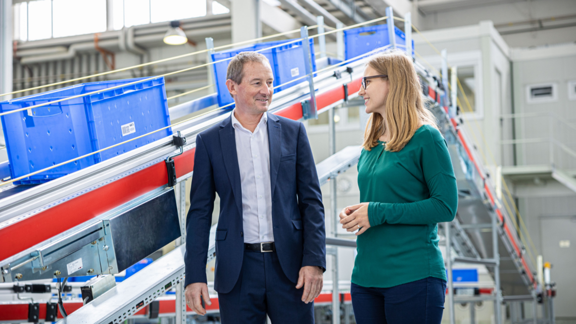 Two employees walking in conversation in front of a conveyor belt with blue crates on top of it
