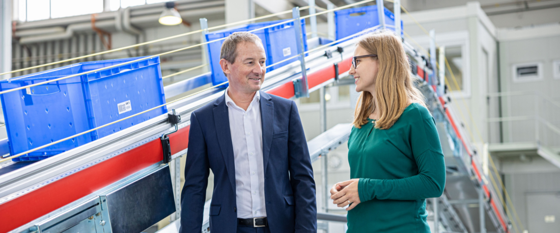 Two employees walking in conversation in front of a conveyor belt with blue crates on top of it