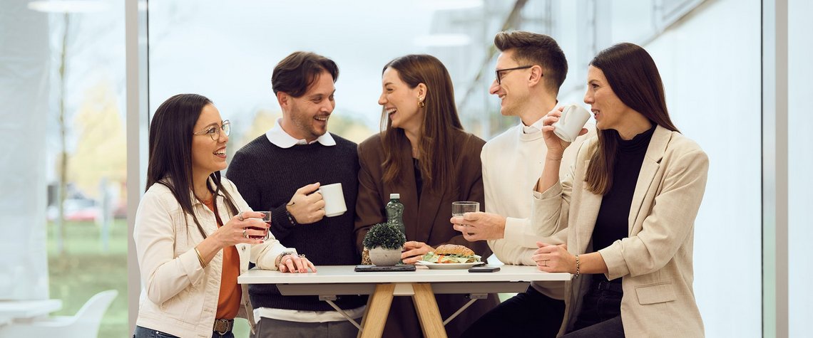 Five TGW Logistics employees laughing at a standing table during a team coffee break