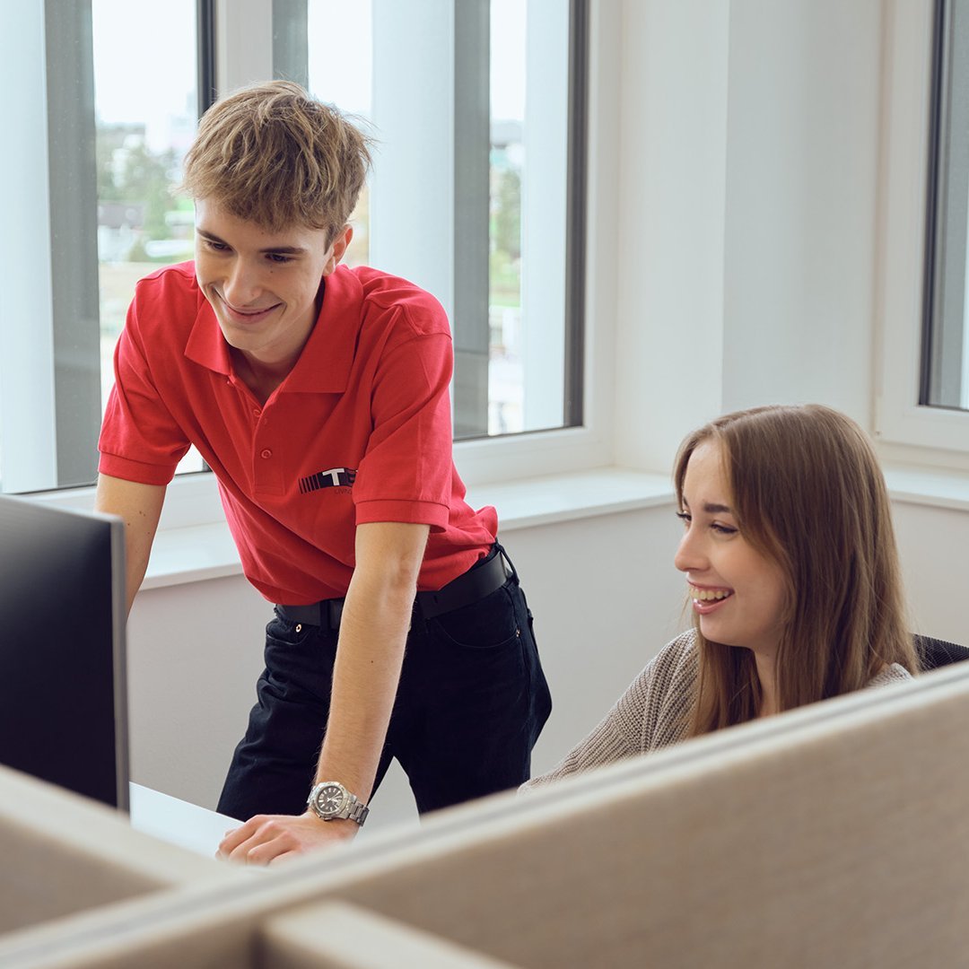 A TGW Logistics apprentice looking at a desktop screen while receiving information from a TGW employee at a desk